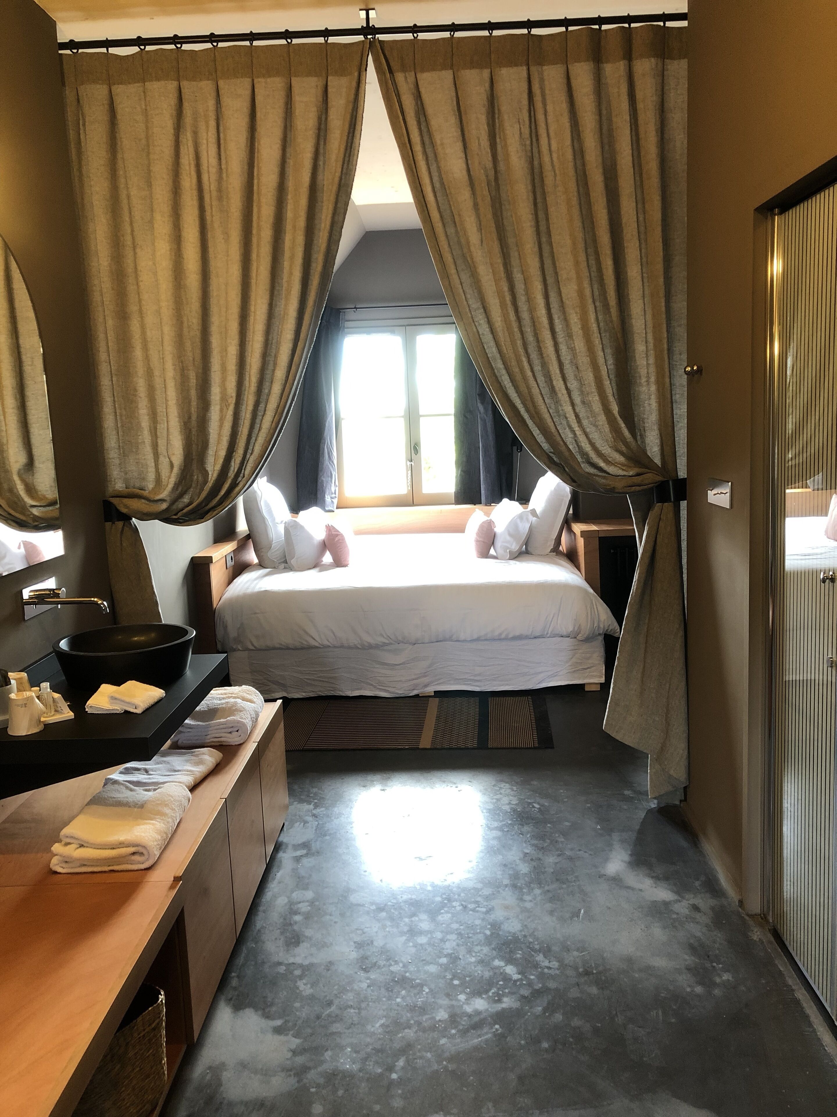 A modern hotel room with a large bed, a bedside table with a black bowl sink, and a wooden bench with towels.
