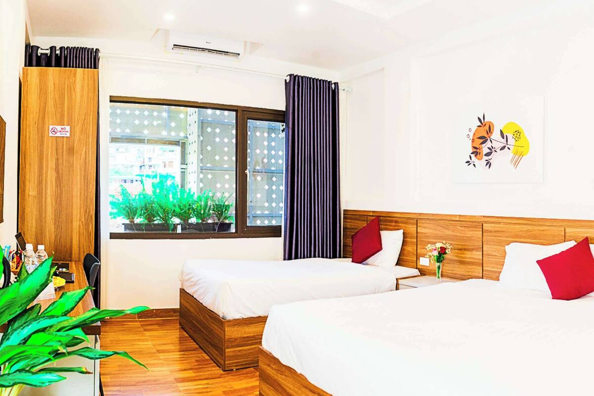 A hotel room with two beds, a wooden headboard, a window with curtains, a wall-mounted air conditioner, and a small table with a plant.