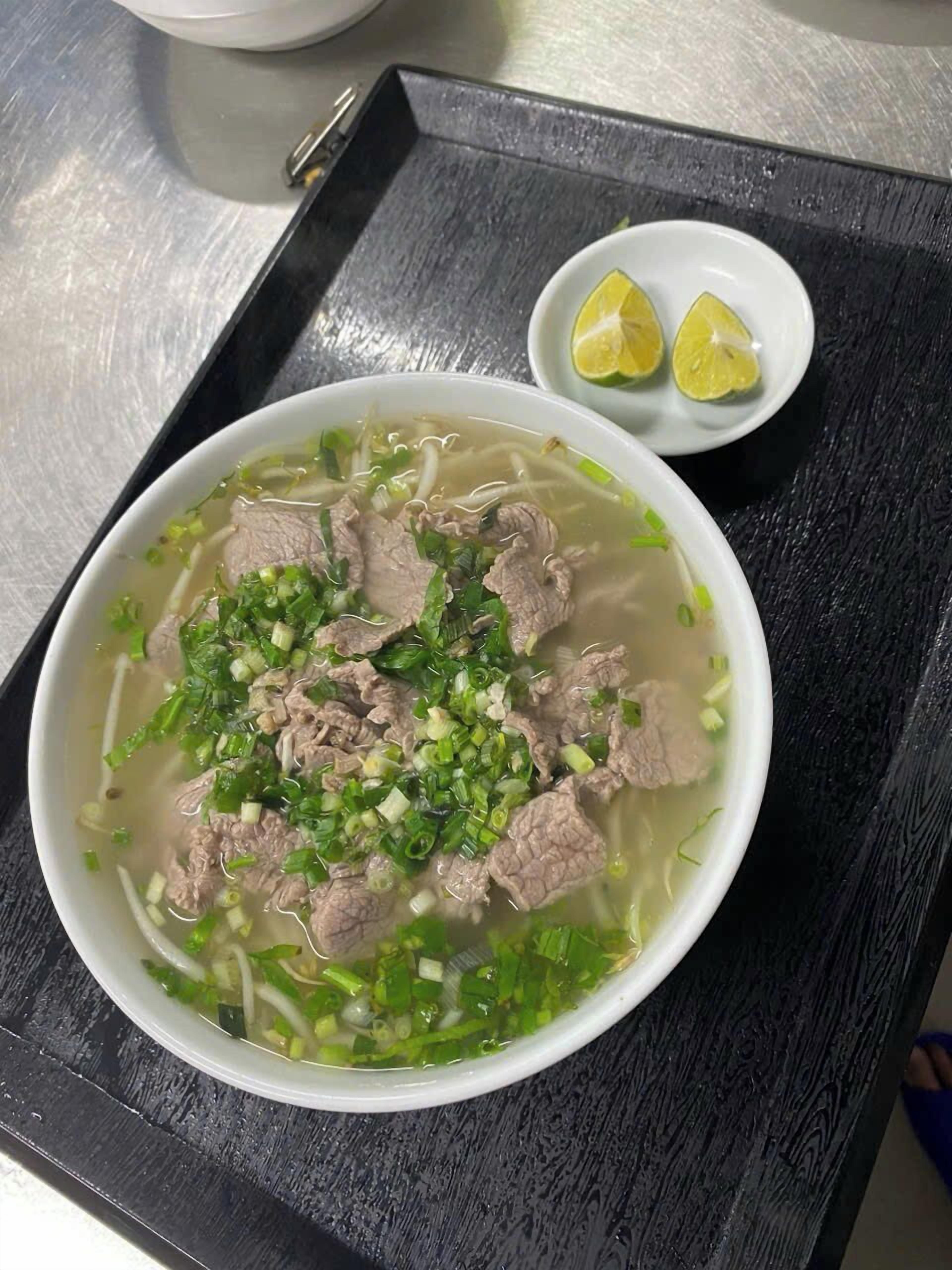 A bowl of beef noodle soup with green onions and lime wedges on a black tray.