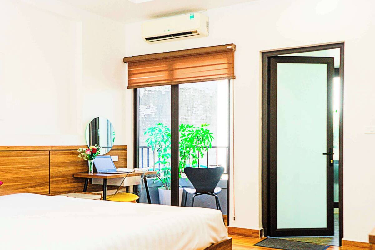 A hotel room with a bed, a desk with a laptop, a chair, a potted plant, and a sliding glass door.