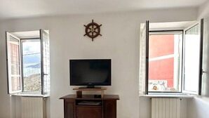Une pièce avec un téléviseur posé sur un meuble en bois, une table en verre et deux fenêtres dont les volets sont ouverts.