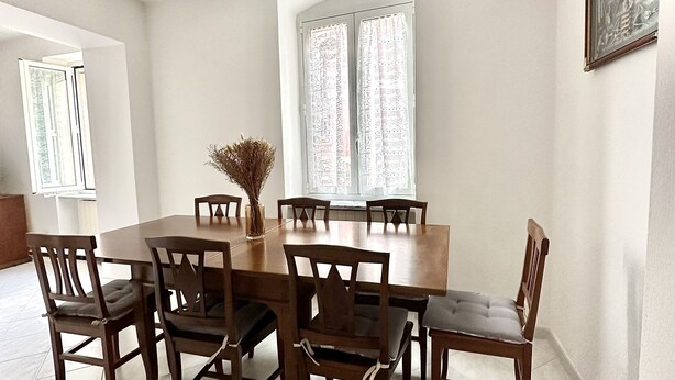 Un coin repas avec une grande table en bois et six chaises, un vase contenant des fleurs séchées et une fenêtre ornée de rideaux à motifs.