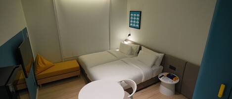 Habitación de hotel pequeña con cama, una mesita, una silla y una mesita de noche con lámpara.