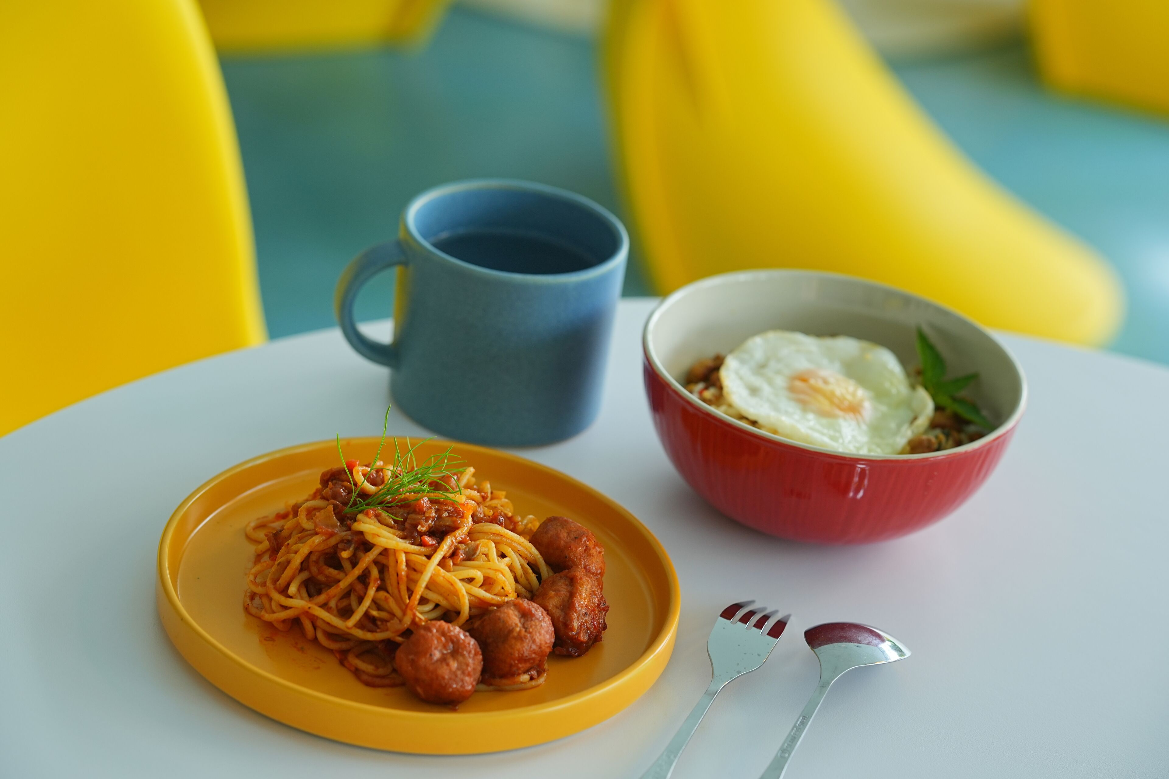 Una mesa con un plato amarillo de espaguetis con albóndigas, un bol rojo con un huevo frito, una taza azul y un tenedor y cuchara.