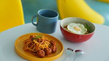 Una mesa con un plato amarillo de espaguetis con albóndigas, un bol rojo con un huevo frito, una taza azul y un tenedor y cuchara.