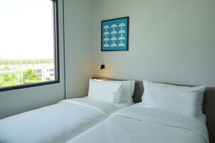 Una habitación de hotel con cama, mesita de noche, un cuadro en la pared y ventana con vistas a edificios y árboles.