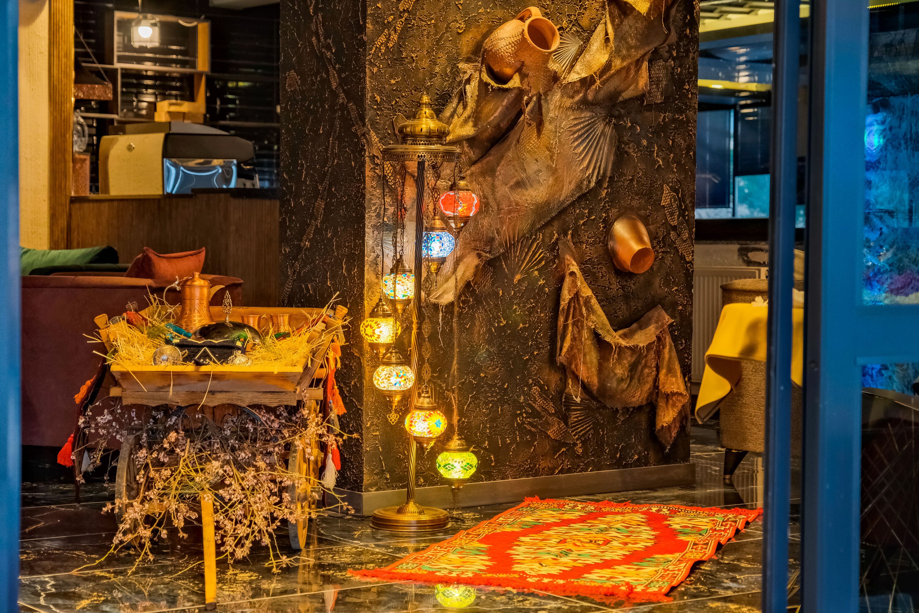 A room with a dark marble floor, a wooden table with a yellow cloth, a red patterned rug, and a wall with a large fish mural and hanging pottery.