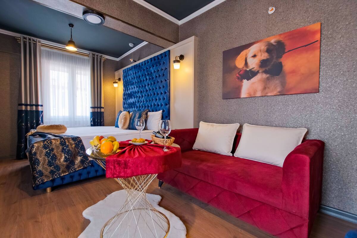 A modern hotel room with a bed, a red sofa, a dining table with fruit and wine, and a painting of a dog.