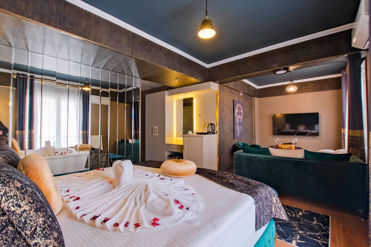 A hotel room with a bed, a sofa, a TV, a mirror, and a kitchenette.