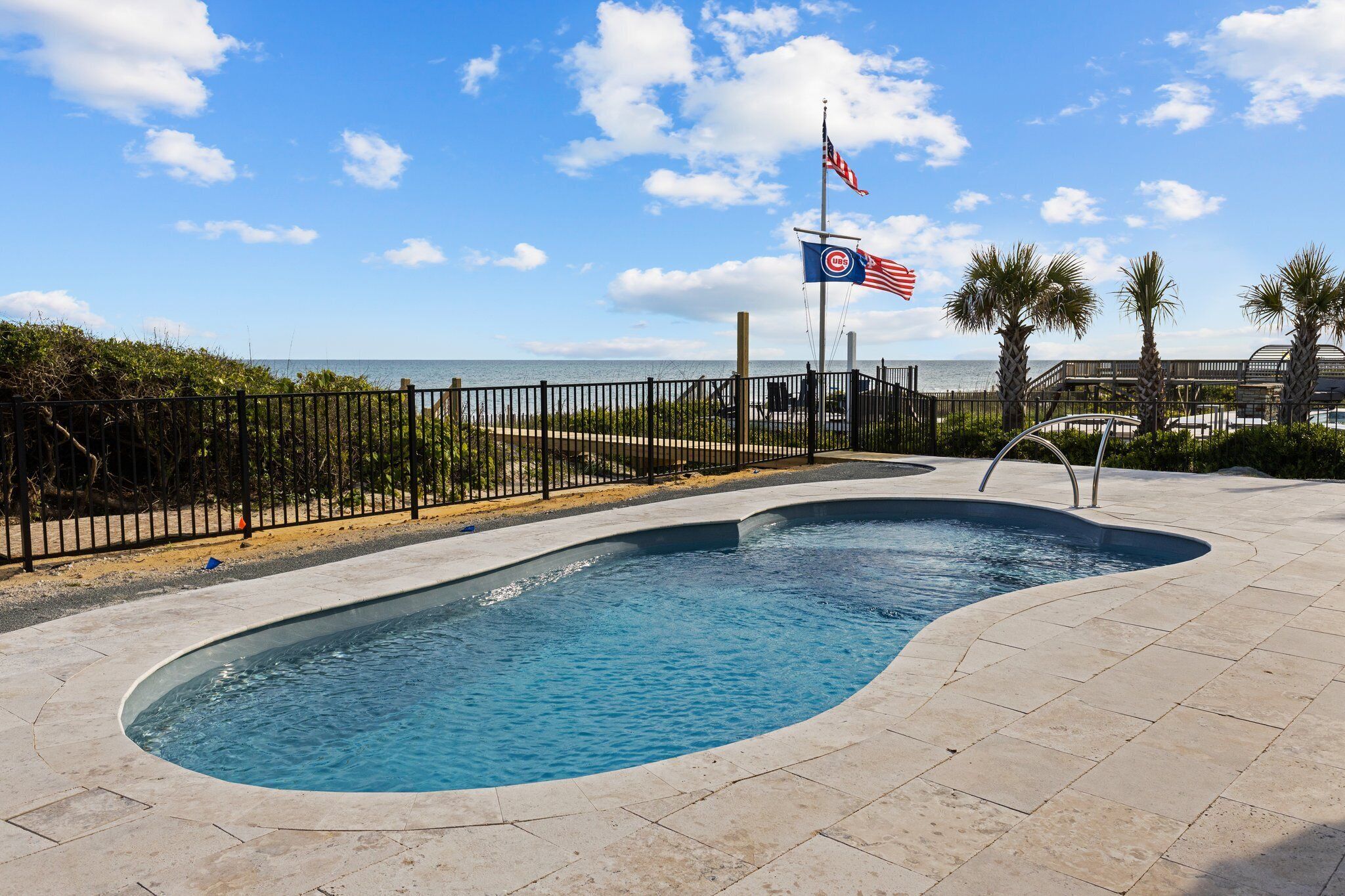 A swimming pool with a curved edge, surrounded by a black metal fence, and a flagpole with an American flag and a Cubs flag.