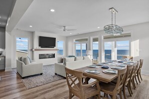 A spacious living room with a large wooden dining table, a fireplace, and a view of the ocean. - Modern 5BR Ocean Front Home w/ Pool! (Emerald Isle)