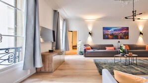 A modern living room with a sofa, a coffee table, and a TV. There is a painting on the wall and a window with curtains. - IN AN ELEGANT SETTING AND FLOODED WITH LIGHT – CHAMPS ELYSEES -RUE JEAN MERMOZ (Paris)