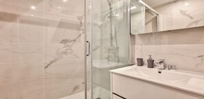Et moderne badeværelse med glasbrusekabine, marmortapetsering, en hvid vask og et spejl over skabet.