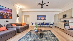 A modern living room with a sofa, coffee table, and a dining area with chairs. There is a kitchen area with cabinets and a microwave. - IN AN ELEGANT SETTING AND FLOODED WITH LIGHT – CHAMPS ELYSEES -RUE JEAN MERMOZ (Paris)