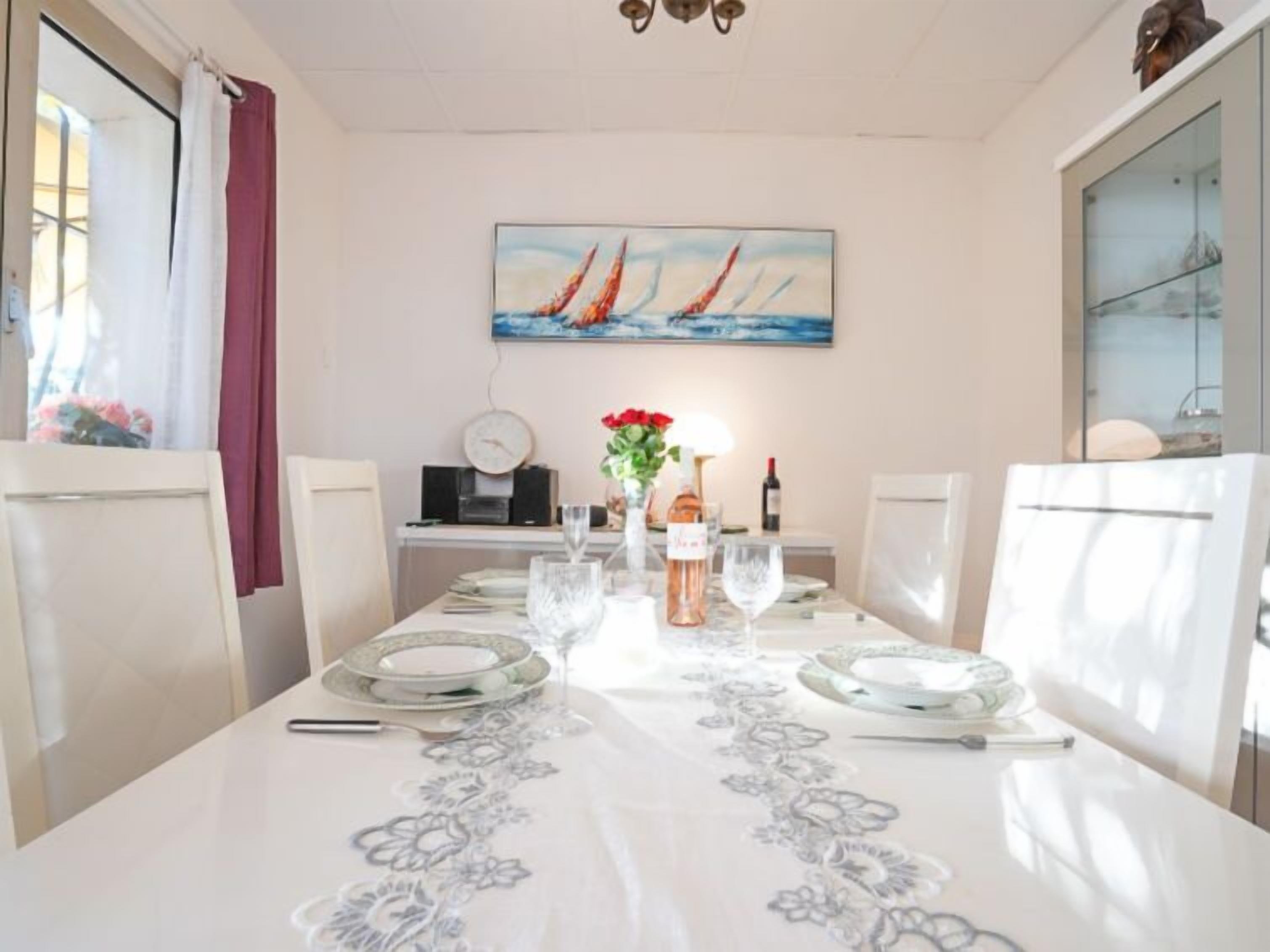 Un comedor con una mesa dispuesta para dos, con mantel de flores, un jarrón de flores rojas y un reloj en la pared.