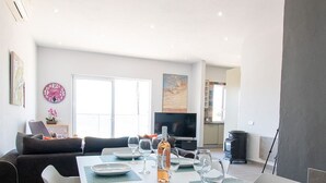 Un coin repas avec une table blanche dressée pour quatre personnes, entourée de chaises blanches dotées de coussins bleus. Un téléviseur à écran plat est fixé au mur au fond de la pièce.