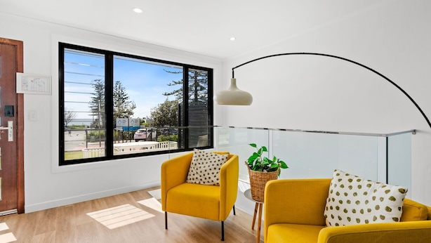 A modern living room with two yellow armchairs, a large window with a view of trees and a parking lot, and a hanging light fixture.