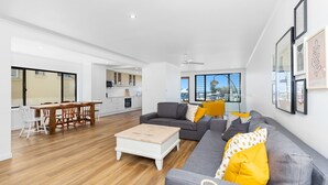 A modern living room with a grey sofa, wooden coffee table, and yellow cushions. There is a dining area with a wooden table and chairs, and a kitchen in the background.