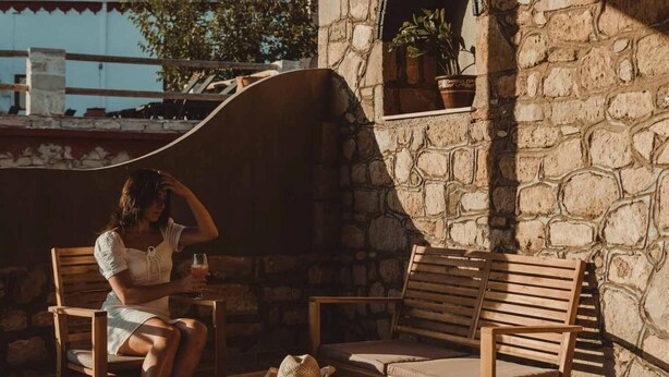 Un edificio de piedra con un banco de madera y una mesa, una mujer sentada en una silla y una planta en maceta.