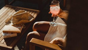 Una silla de madera con cojín, un vaso de bebida rosa y una cesta de libros sobre una mesa de madera.