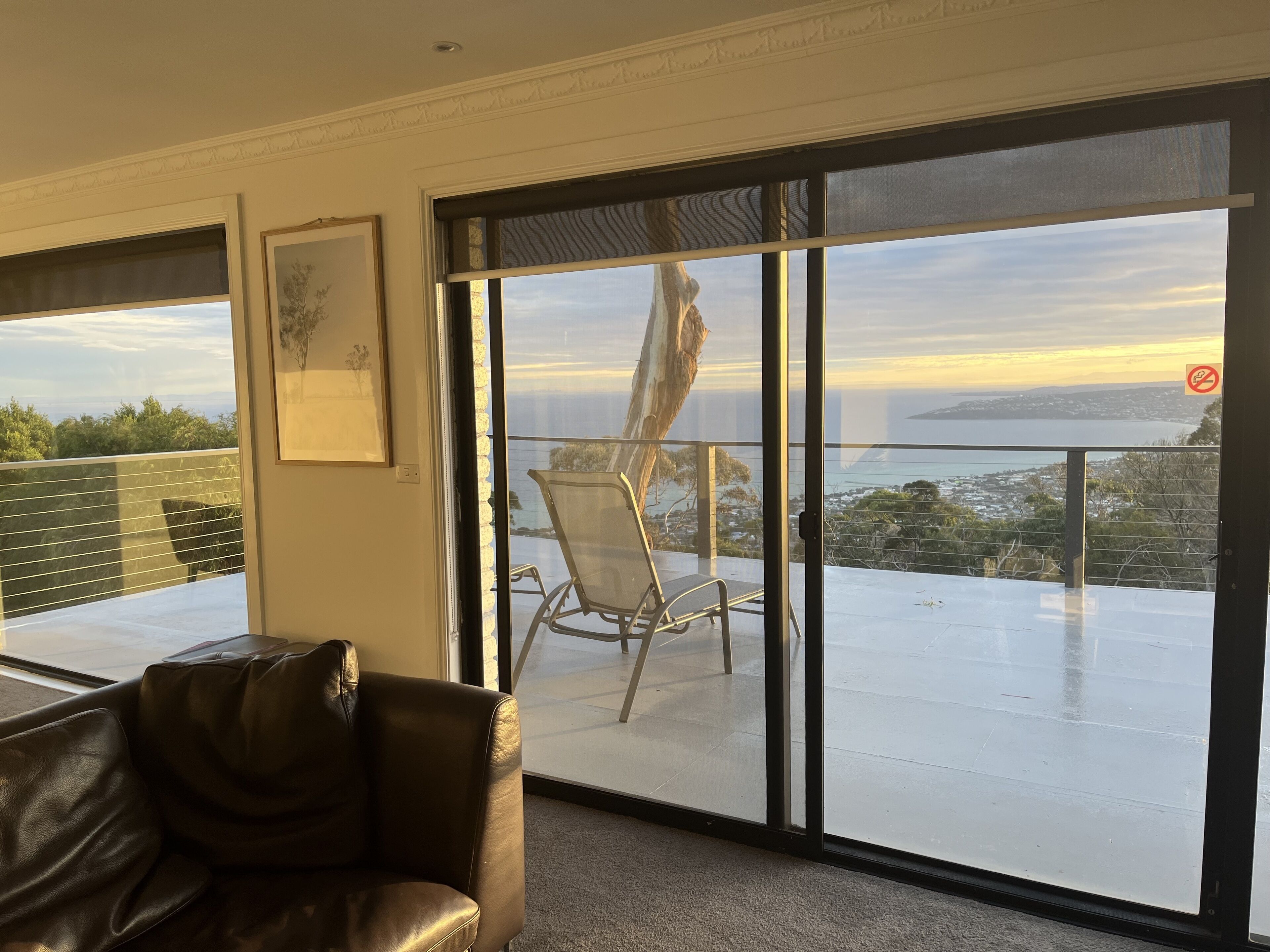 A room with a large sliding glass door leading to an outdoor balcony with a view of the sea and a sunset. There is a leather sofa and a framed picture on the wall.