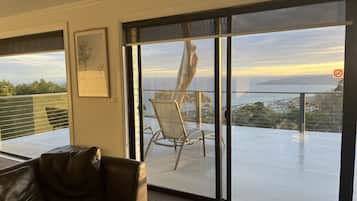 A room with a large sliding glass door leading to an outdoor balcony with a view of the sea and a sunset. There is a leather sofa and a framed picture on the wall.