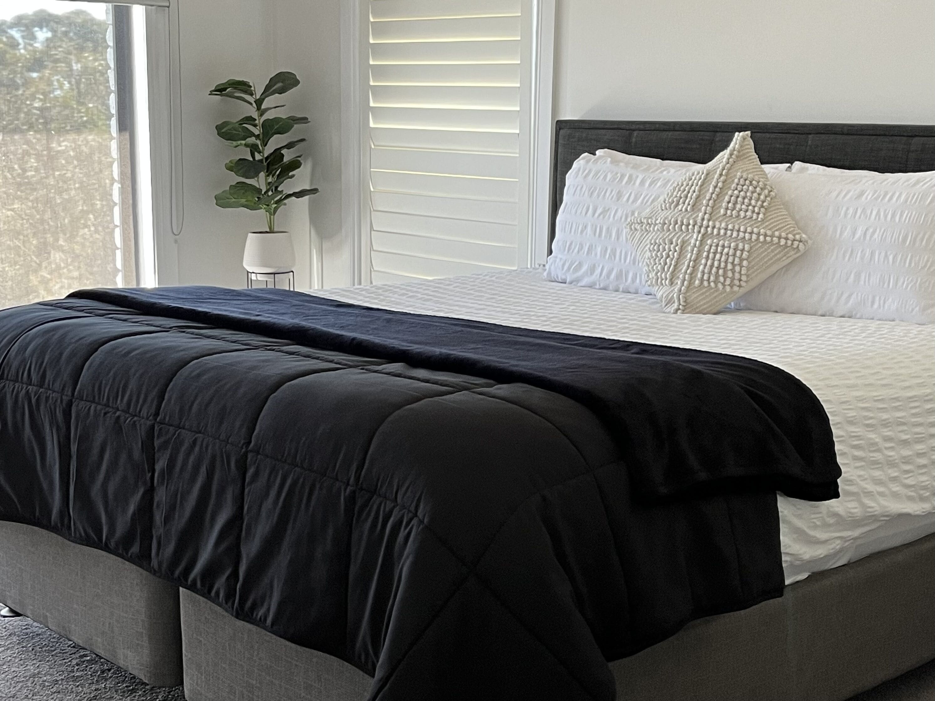 A bedroom with a bed, a window with blinds, a potted plant, and a bedside table.