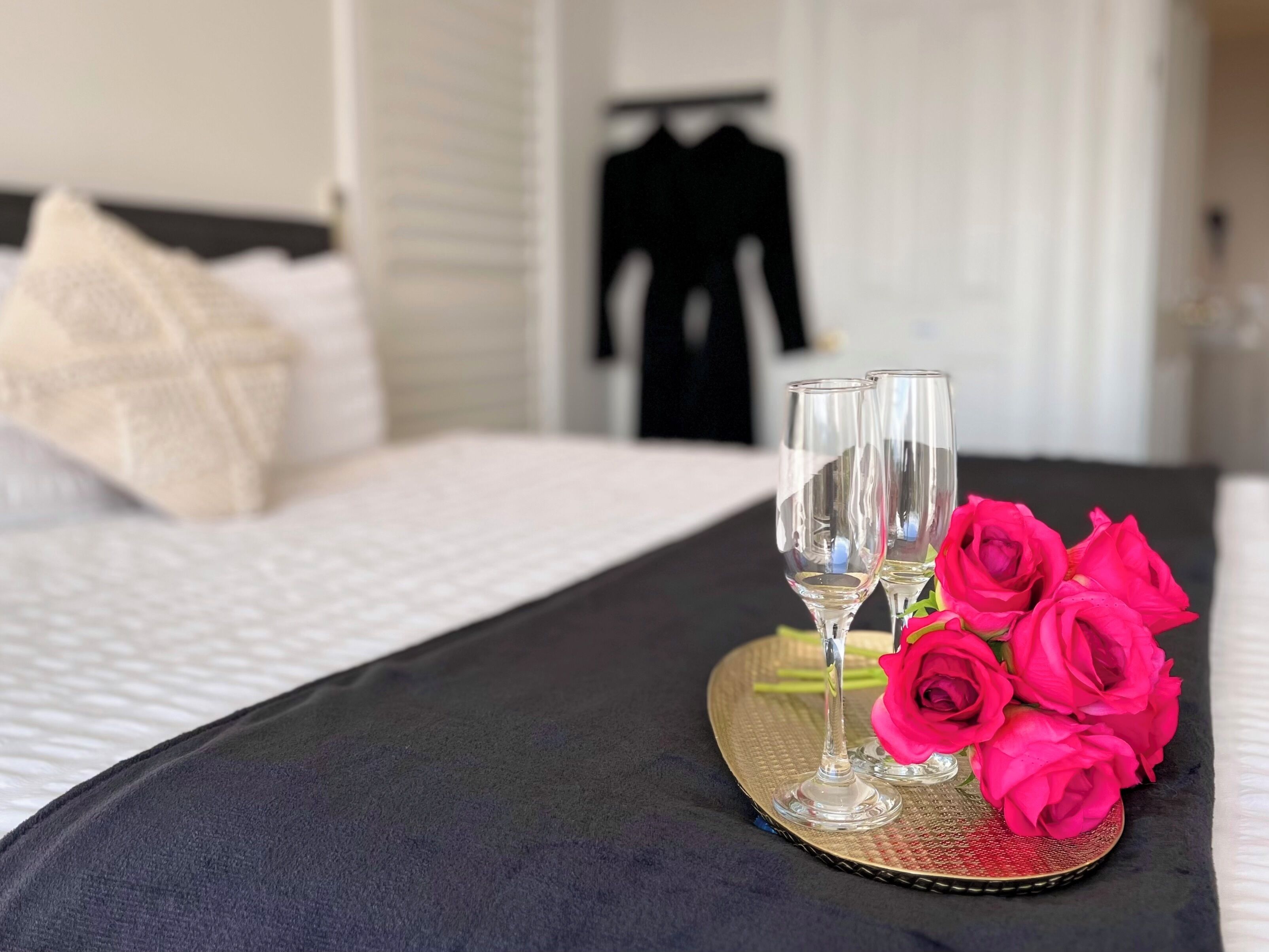 Una cama bien tendida con un edredón negro, dos copas de vino vacías y un ramo de rosas rosadas sobre una bandeja de mimbre.