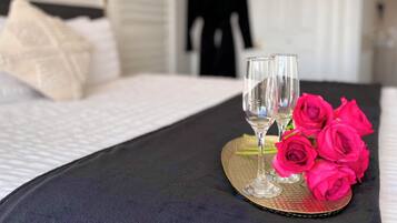 Una cama bien tendida con un edredón negro, dos copas de vino vacías y un ramo de rosas rosadas sobre una bandeja de mimbre.