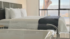 A modern hotel room with a large bathtub, a neatly made bed with white bedding, and a view of a tree through a large window. - Arthurs Holiday Villa (Arthurs Seat)