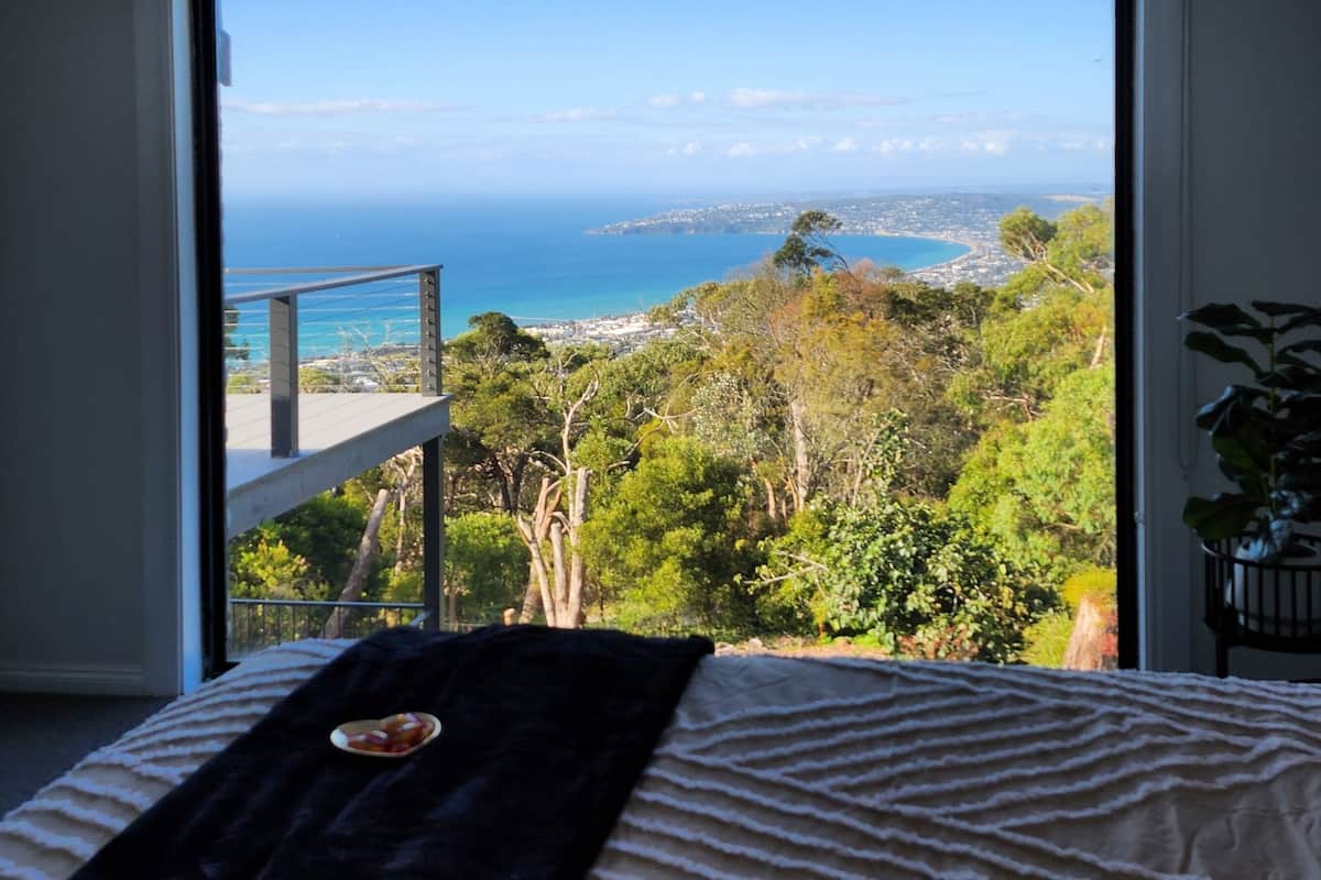 Une chambre avec une grande fenêtre donnant sur un paysage côtier, un lit recouvert d’un jeté noir et une vue sur un balcon.
