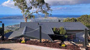 A house with a sloped roof, a large tree in front, and a view of a coastal town and ocean. - Arthurs Holiday Villa (Arthurs Seat)
