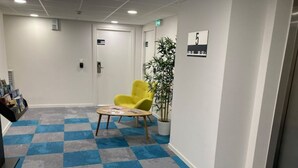 Aula, jossa on ruutukuviollinen matto, keltainen tuoli, vastaanottotiski ja ruukussa oleva kasvi.