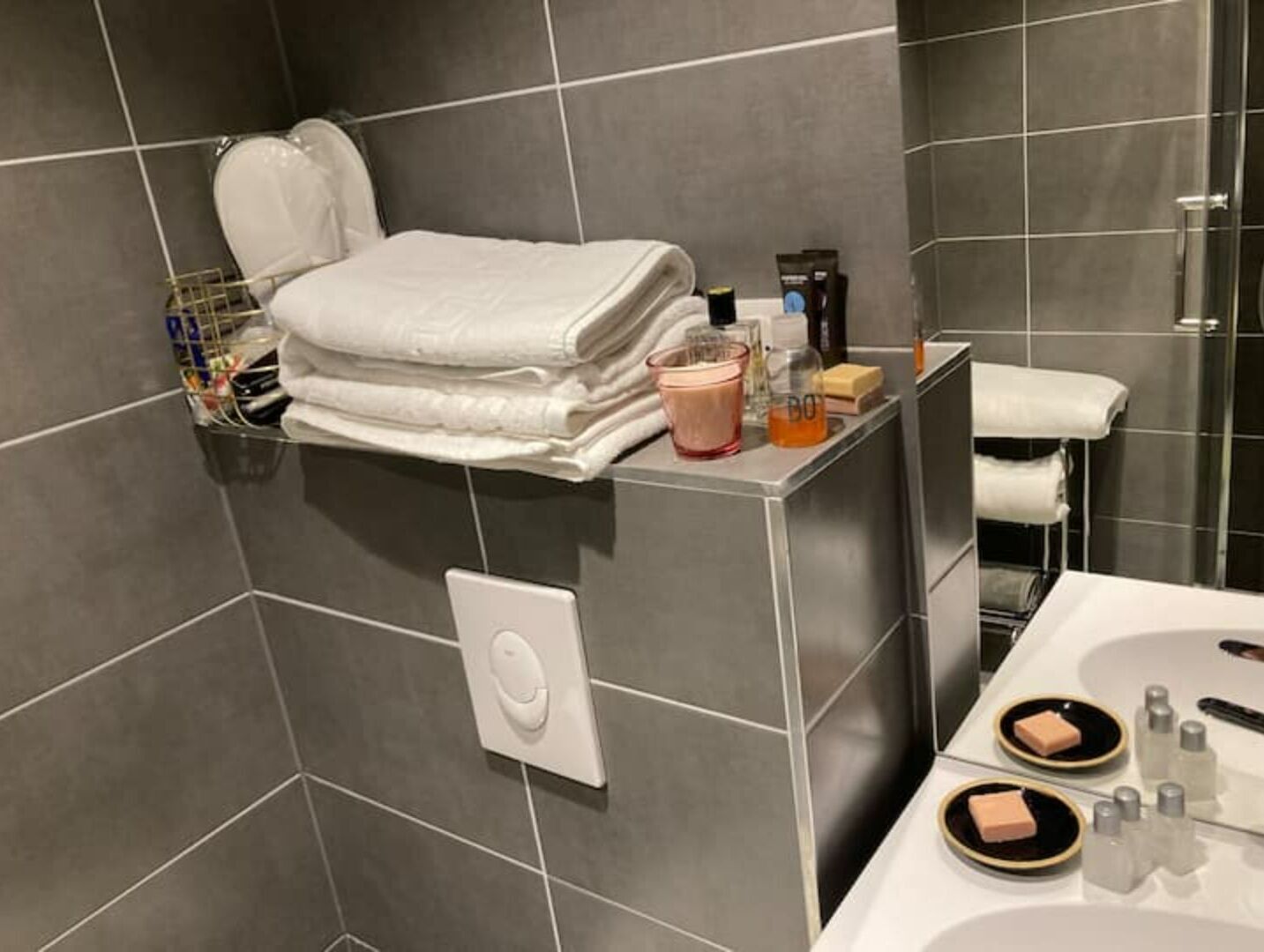Une salle de bain avec des carreaux gris, une serviette blanche posée sur une étagère, des articles de toilette et un lavabo blanc avec du savon et de la lotion.