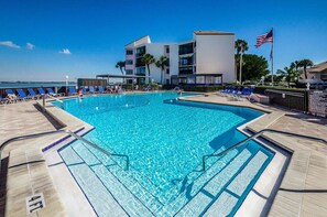A swimming pool with clear blue water, surrounded by lounge chairs and a building with multiple floors and balconies.