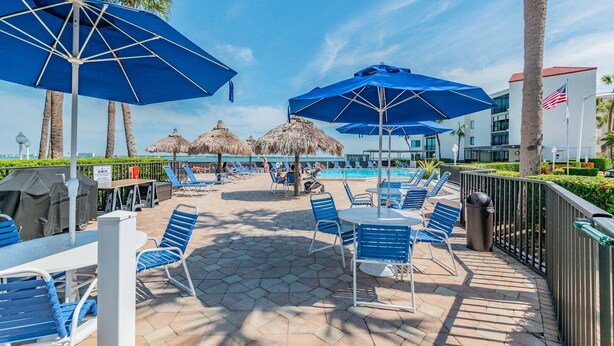 Zona junto a la piscina con sombrillas azules, sillas de descanso y una piscina al fondo.
