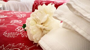 A bedroom with a bed covered in a red floral bedspread, white pillows, and a white bathrobe. There is a large white flower on the bed, and a hanging plant with a wooden hanger. A framed picture of flowers is on the wall. - An exclusive terraced house in a very central position, 600 meters from the sea. (San Benedetto del Tronto)