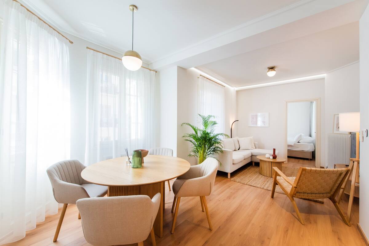 Ein modernes Wohnzimmer mit einem runden Holztisch, beigefarbenen Sesseln, einem weißen Sofa, einer Topfpflanze und einem kleinen Beistelltisch.
