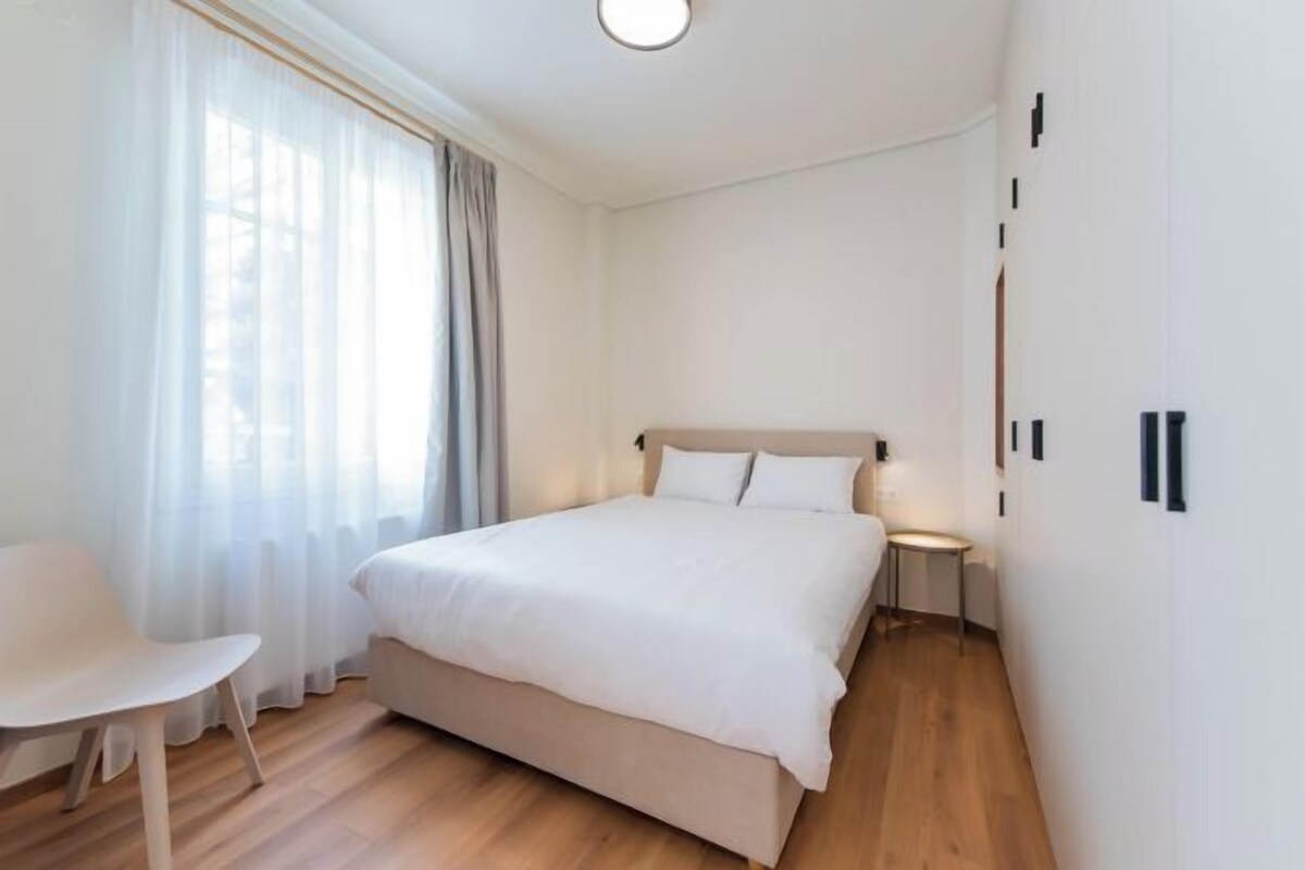 Un dormitorio con cama, una silla, mesita de noche y una ventana con cortinas.