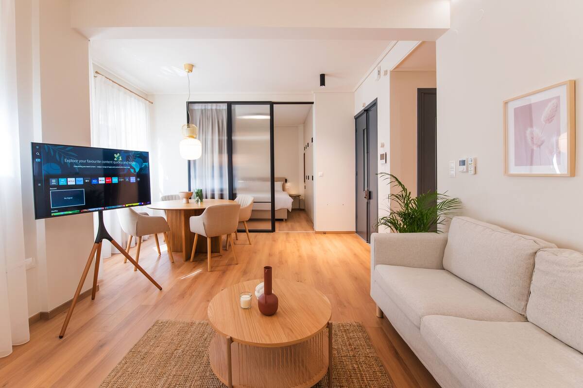 Ein modernes Wohnzimmer mit Flachbildfernseher, rundem Holztisch, beiger Couch und Essbereich mit Tisch und Stühlen.