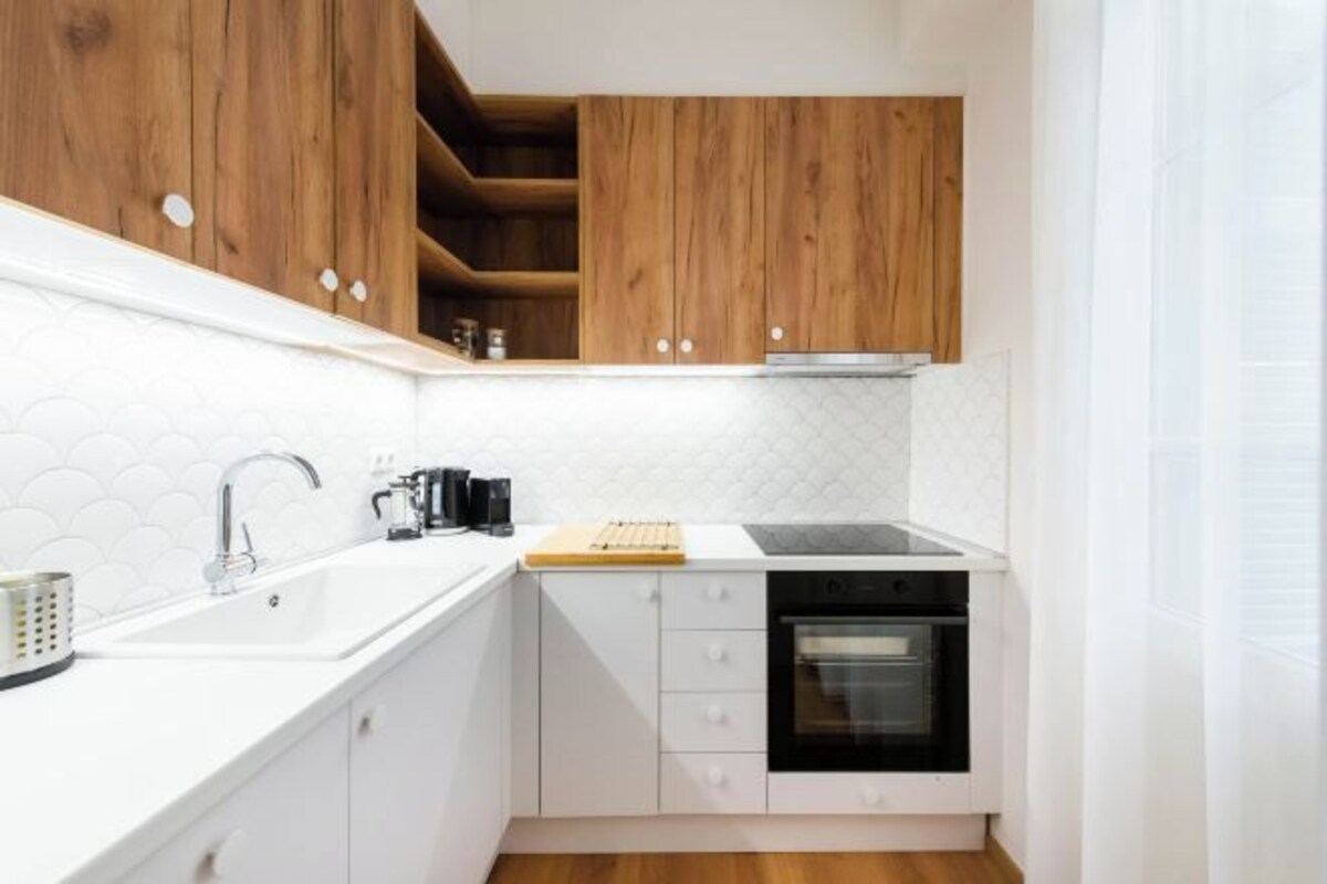 A compact kitchen with white cabinets, a wooden upper shelf, a sink, a stove, and a countertop with a cutting board.