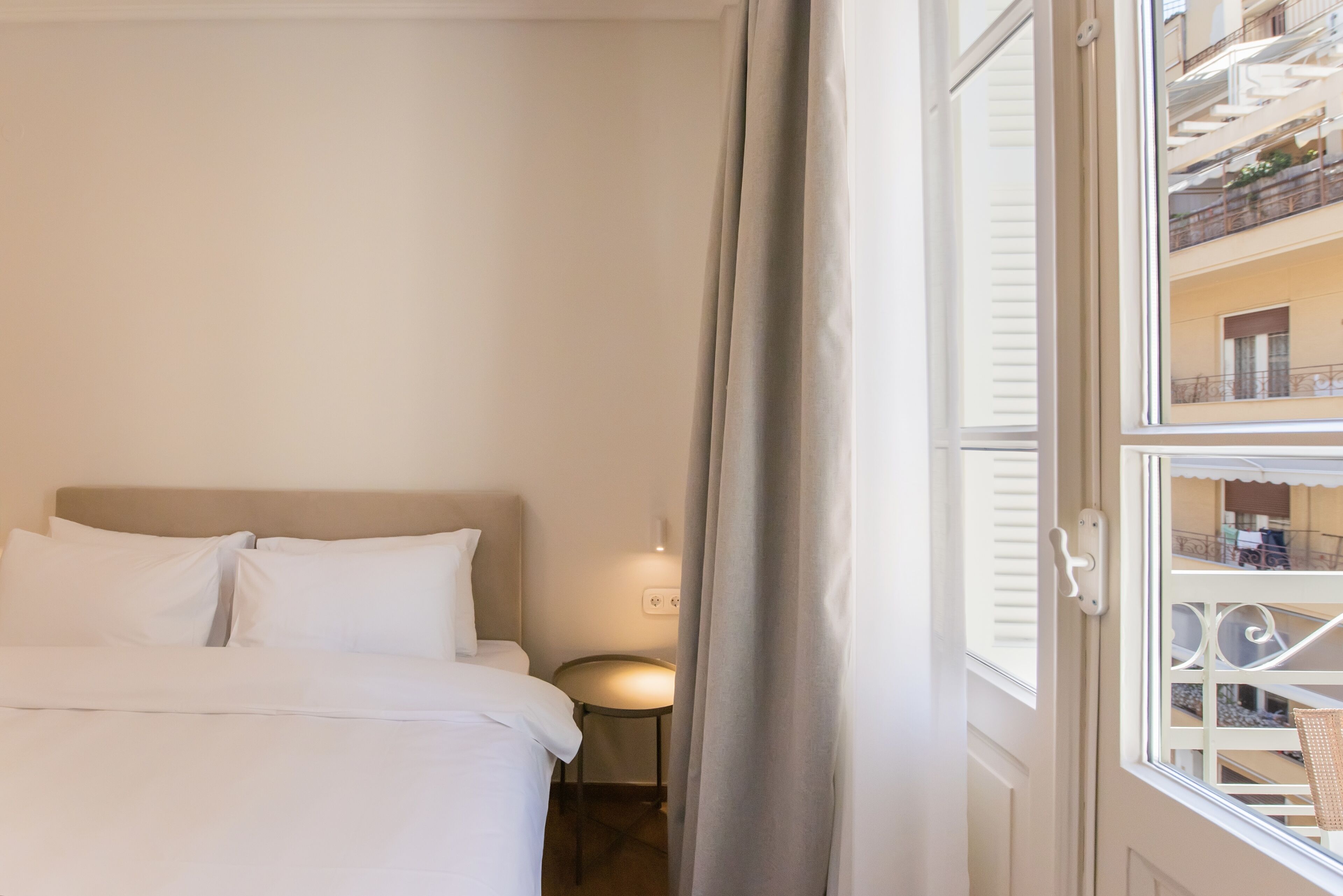 A hotel room with a bed, a nightstand, a lamp, and a view of the building's exterior through a window.