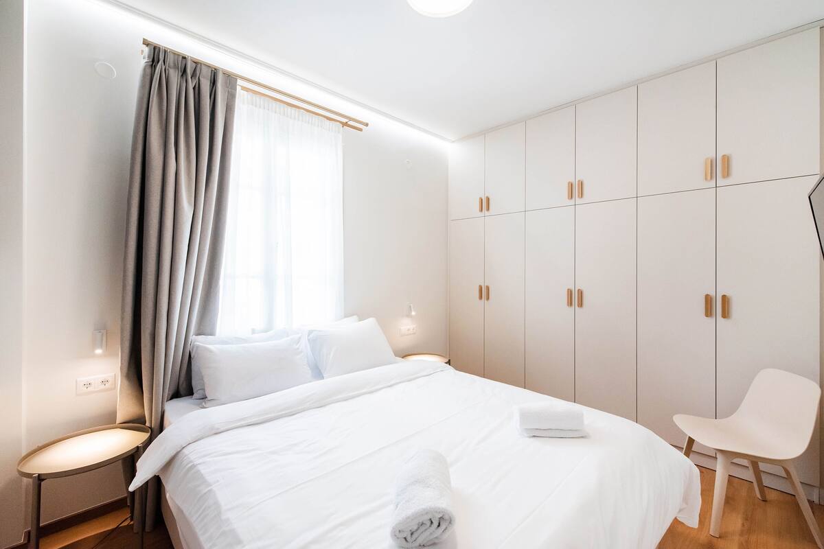 Ein modernes Hotelzimmer mit einem ordentlich bezogenem Bett, einem Sessel und einem Kleiderschrank mit mehreren Fächern.