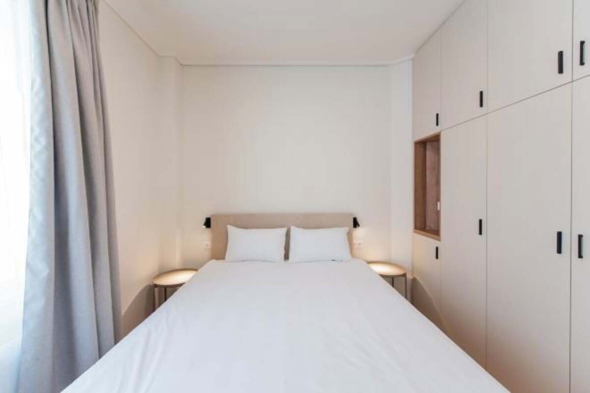 A minimalist bedroom with a large bed, two bedside lamps, and a wardrobe with multiple doors.