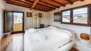Un dormitorio con techo de madera, una cama con ropa de cama blanca, una mesita de noche con lámpara y vistas de la ciudad a través de amplios ventanales.