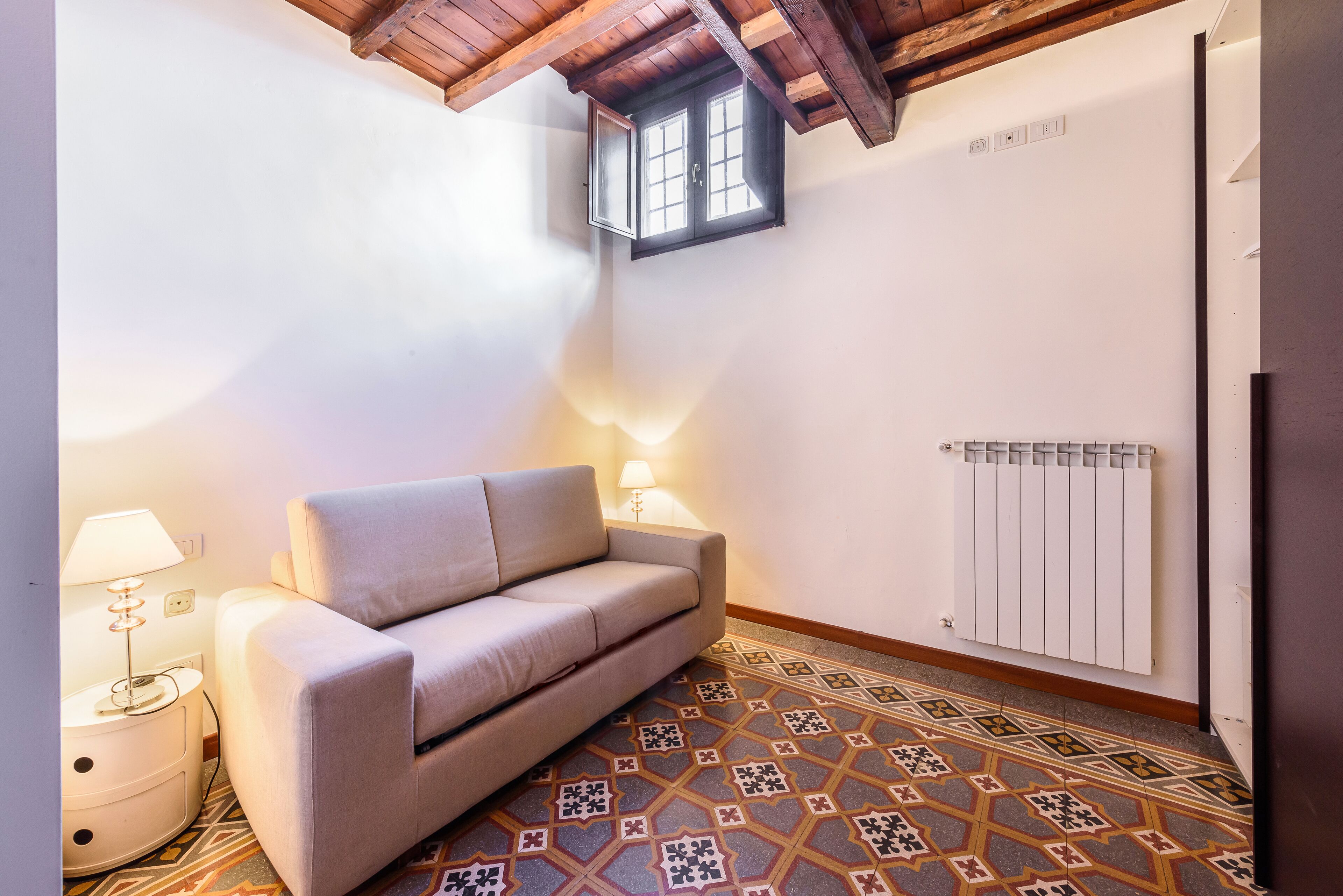 A cozy room with a beige sofa, a patterned floor, a radiator, and a window with a view.