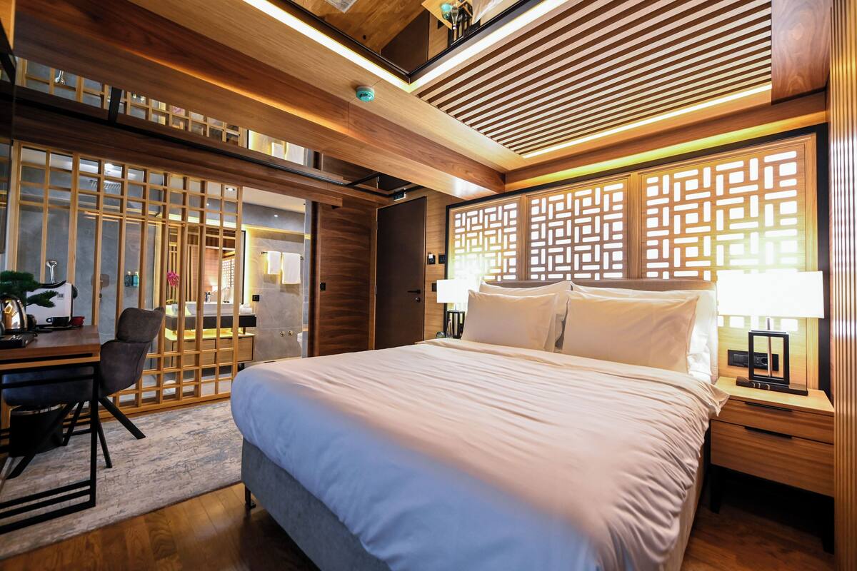 A modern hotel room with a large bed, a desk with a chair, and a wooden partition with intricate patterns.