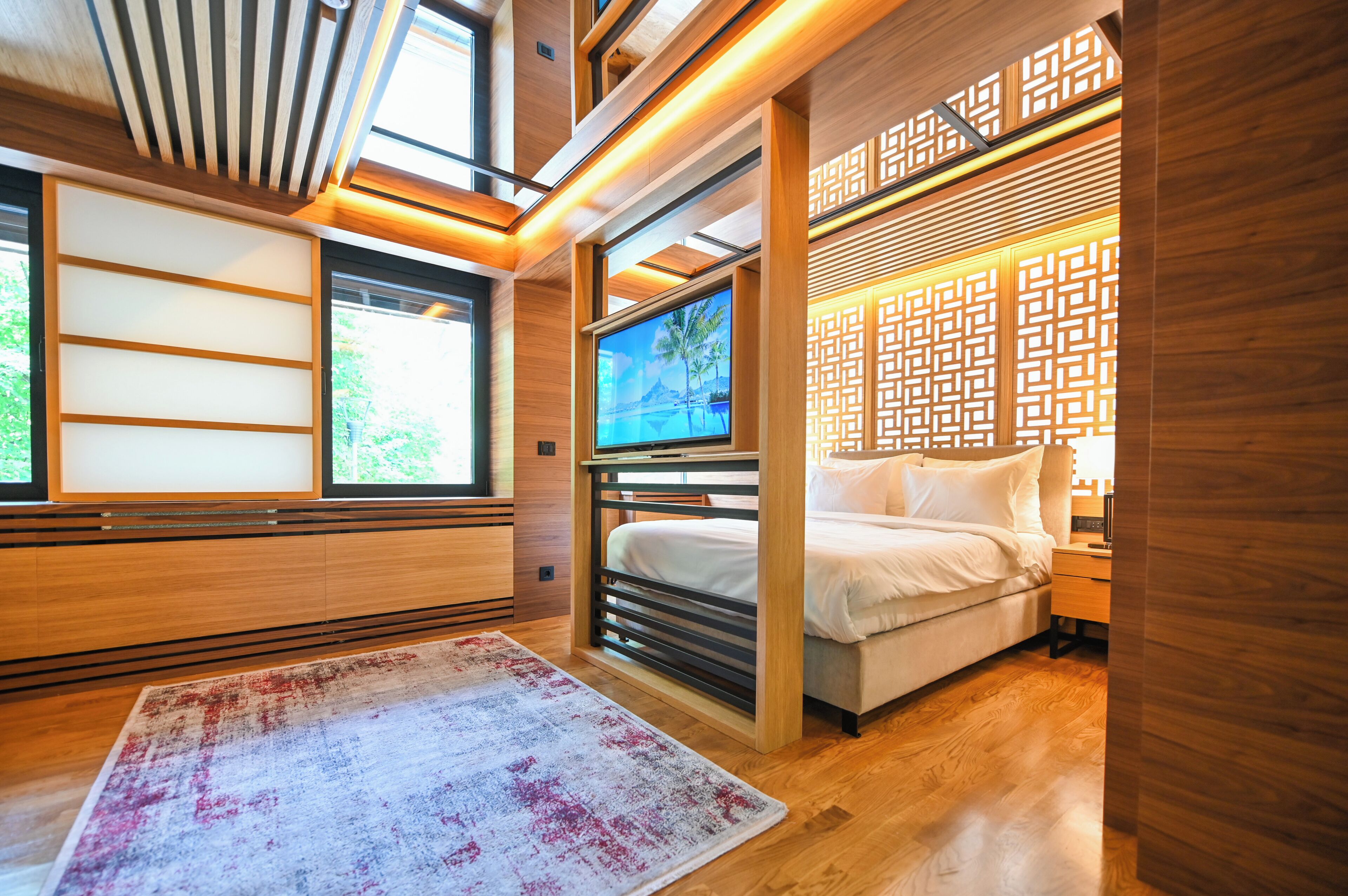 Een moderne slaapkamer met een bed, een televisie en uitzicht op groen door grote ramen.