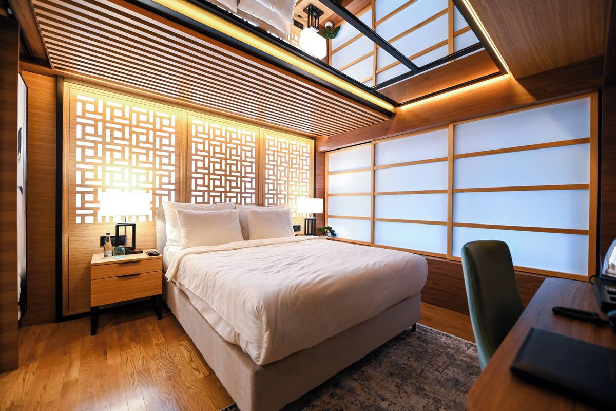 A modern hotel room with a large bed, wooden headboard, and a view of the city through large windows.