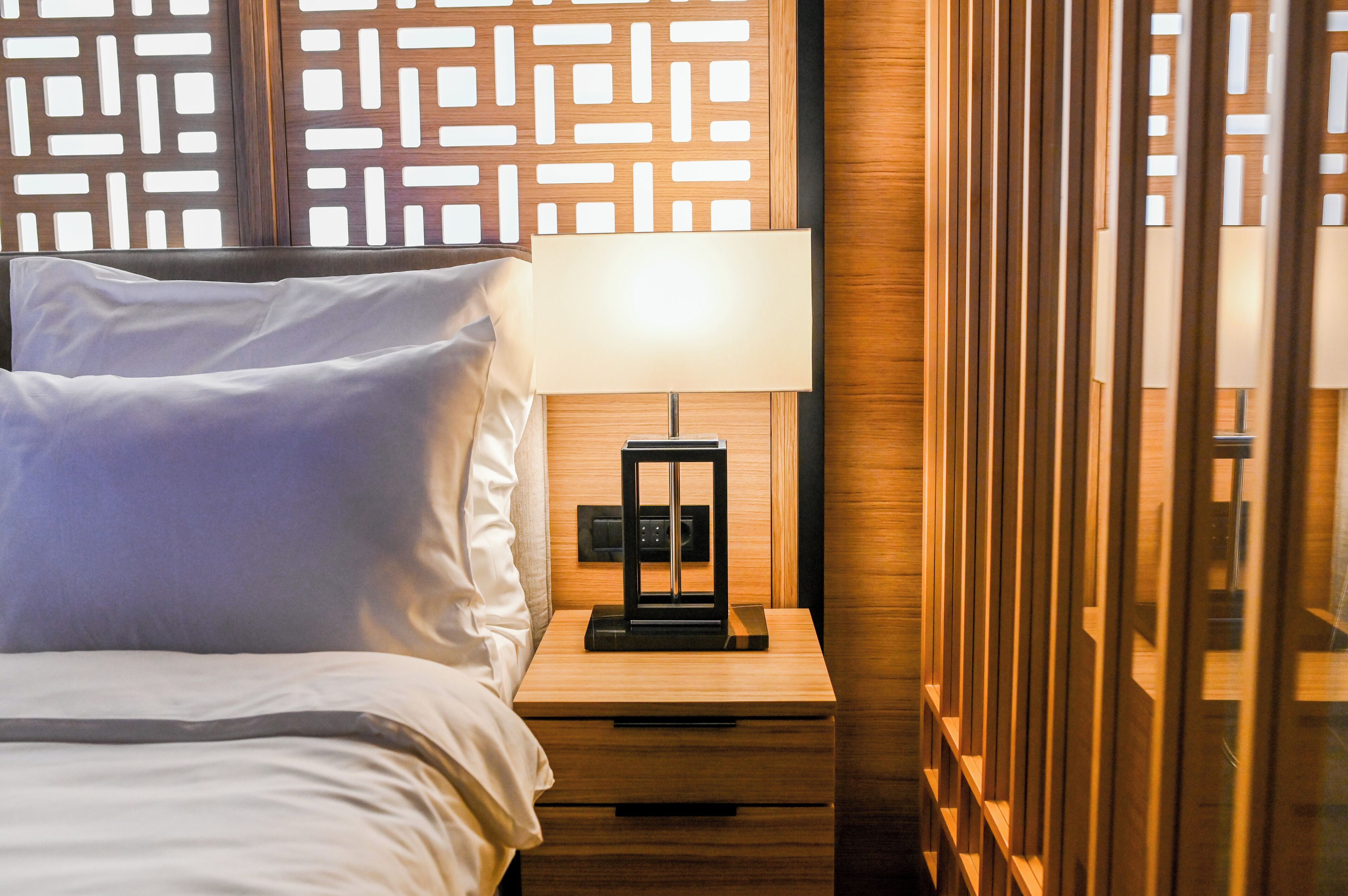 A neatly made bed with white linens, a bedside table with a lamp, and a wooden panel wall with a geometric pattern.